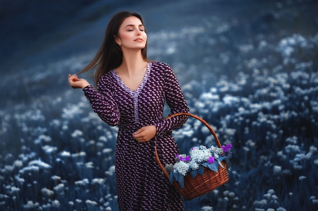 Une belle femme vêtue d'une robe d'été dans un champ de fleurs épanouies avec un panier de paille dans les mains