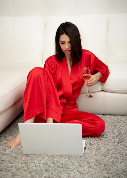 Une belle femme vêtue d'une robe rouge est assise sur le sol et boit du vin travaille devant un ordinateur