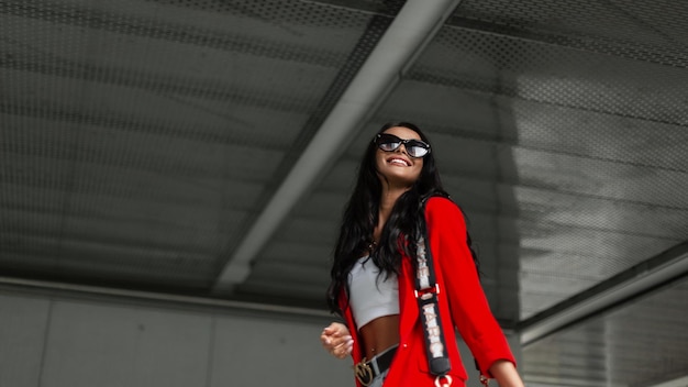 Belle fille d'affaires heureuse à la mode avec un sourire dans des vêtements décontractés lumineux à la mode avec un haut blazer rouge et un sac marchant et s'amusant dans la ville