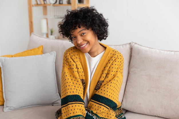 Belle fille afro-américaine avec une coiffure afro souriante assise sur un canapé à la maison intérieure jeune afr