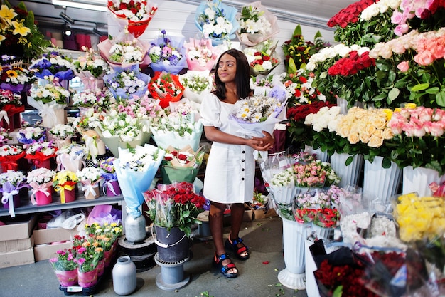 Belle fille afro-américaine en robe blanche tendre avec bouquet de fleurs dans les mains debout sur fond floral dans le magasin de fleurs. Fleuriste femme noire.