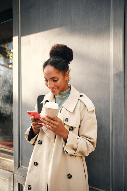 Belle fille afro-américaine souriante en trench-coat élégant avec du café pour aller joyeusement en utilisant un téléphone portable en plein air