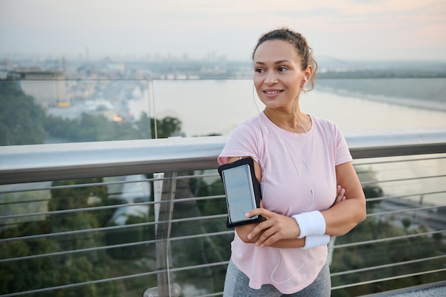 Belle fille afro-américaine sportive et heureuse avec des écouteurs debout avec les bras croisés sur le pont de la ville et un sourire à pleines dents souriant en détournant les yeux. Femme athlète se reposant après un entraînement cardio intense à l'extérieur