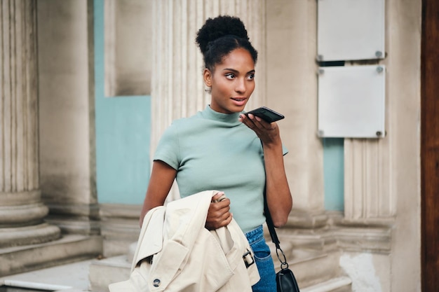 Belle fille afro-américaine avec trench-coat à la main parlant pensivement sur téléphone portable dans la rue de la ville