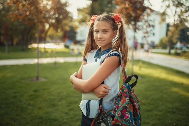 Une belle fille d'âge scolaire est debout près d'un arbre et tient une tablette. Écolière portant une jupe et une chemise avec des chaussures