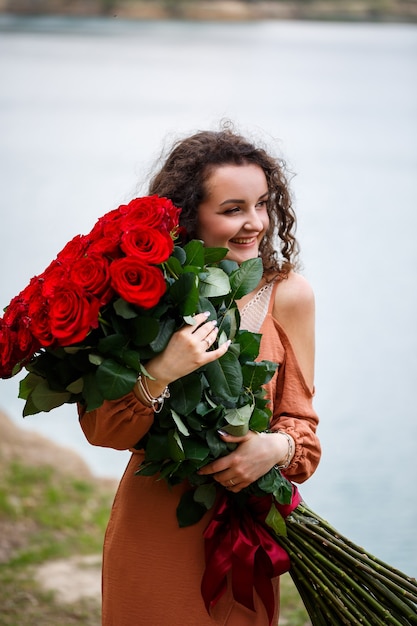 Une belle fille d'apparence européenne aux cheveux bouclés et un sourire sur son visage avec un énorme bouquet de roses rouges sur fond de lac bleu. Chaude journée d'été, jeune femme heureuse, émotions de joie