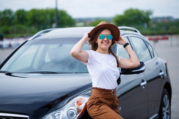 Une belle fille d'apparence européenne avec des lunettes et un chapeau marron se tient près d'une voiture noire. Photo près de la voiture.