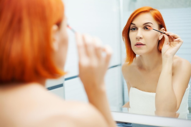 Belle fille applique des cosmétiques sur le visage dans une salle de bain lumineuse