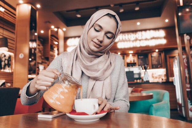 Belle fille arabe avec un foulard sur la tête dîne dans un café confortable, boit du thé à l'argousier