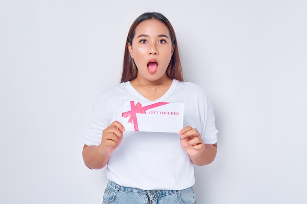 Belle fille asiatique choquée portant un t-shirt blanc décontracté tenant une carte de bon de coupon de certificat-cadeau en regardant la caméra isolée sur fond blanc Concept de mode de vie des gens