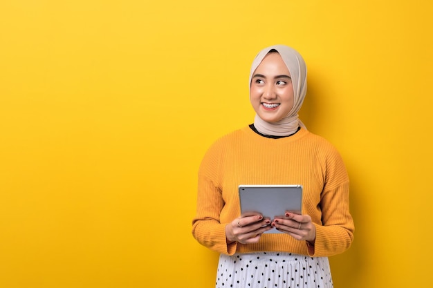 Belle fille asiatique heureuse souriante portant le hijab tenant une tablette numérique et regardant de côté l'espace de copie isolé sur fond jaune