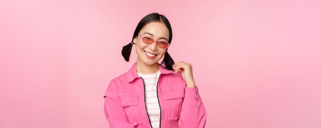 Belle fille asiatique en lunettes de soleil souriant à la caméra posant sur fond de studio rose