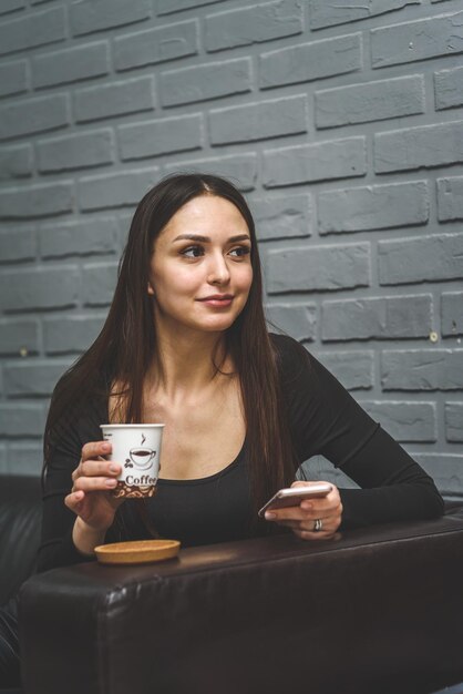 Belle fille assise sur un canapé en cuir dans le salon buvant du café et parlant au téléphone