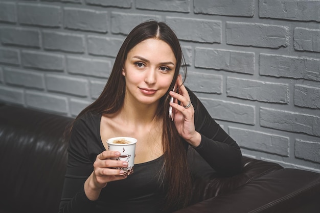 Belle fille assise sur un canapé en cuir dans le salon buvant du café et parlant au téléphone