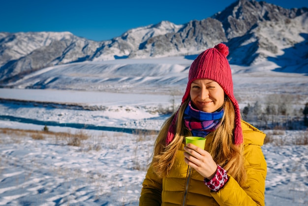 Belle fille au chapeau rouge tient le verre sur le fond des montagnes enneigées par une journée ensoleillée glaciale. Gaie jeune femme profitant des vacances de Noël dans les montagnes, regardant la caméra et souriant.