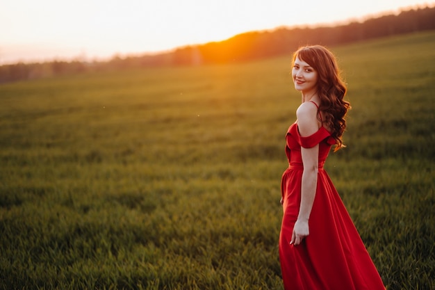 Une belle fille au printemps dans une robe rouge marche dans un champ au coucher du soleil