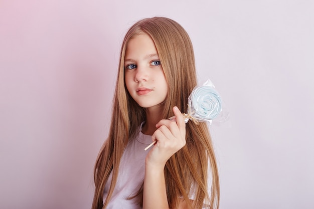 Belle fille aux cheveux blonds mange une sucette, caramel rond sur bâton dans les mains d'une fille souriante gaie. Petite fille aux cheveux longs en t-shirt blanc lèche Lollipop. Russie, Sverdlovsk, 1er décembre 2018