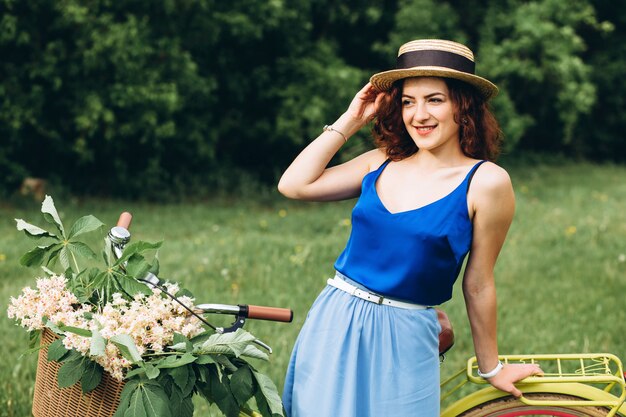 Belle fille aux cheveux bouclés dans un chapeau avec vélo avec panier de fleurs