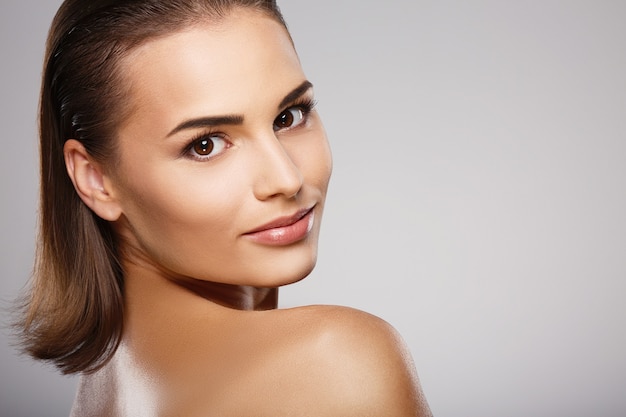 Belle fille aux cheveux bruns, peau fraîche et propre et épaules nues posant au mur gris, un modèle avec un maquillage nu léger, gros plan.