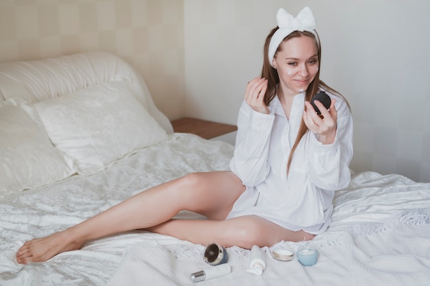 Belle fille aux cheveux longs au lit prend soin de sa peau. Utiliser des crèmes cosmétiques