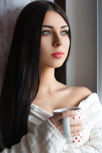 Belle fille aux cheveux longs, boire du café par la fenêtre