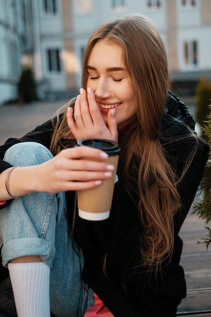 Belle fille aux cheveux longs avec un café assis sur un banc et riant