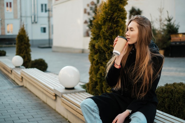 Une belle fille aux cheveux longs avec du café assise sur un banc et regardant au loin