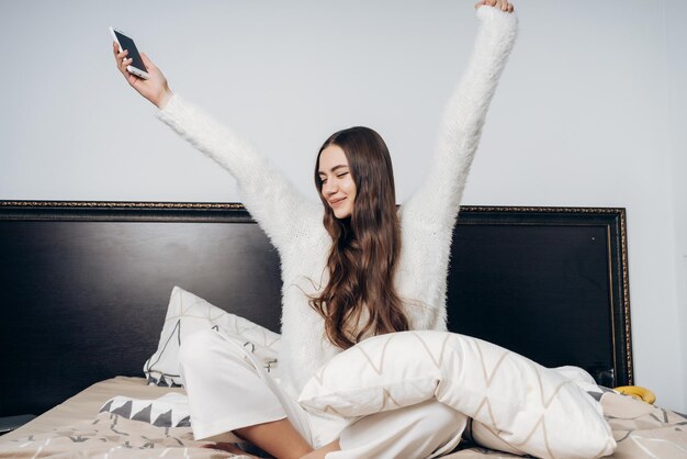 Belle fille aux cheveux longs en pyjama est assise sur le lit et s'étire se réveilla tôt le matin