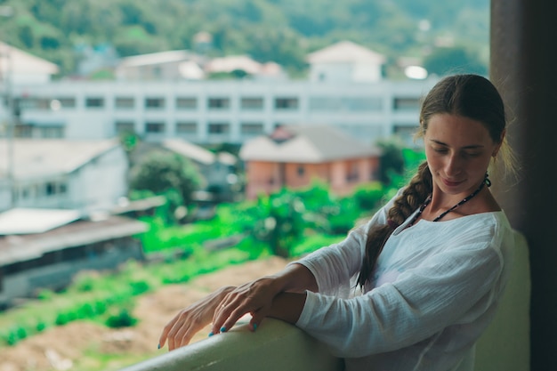 belle fille aux cheveux longs, se prélassant dans la robe blanche sur le balcon de l'hôtel avec vue sur les plantes tropicales.