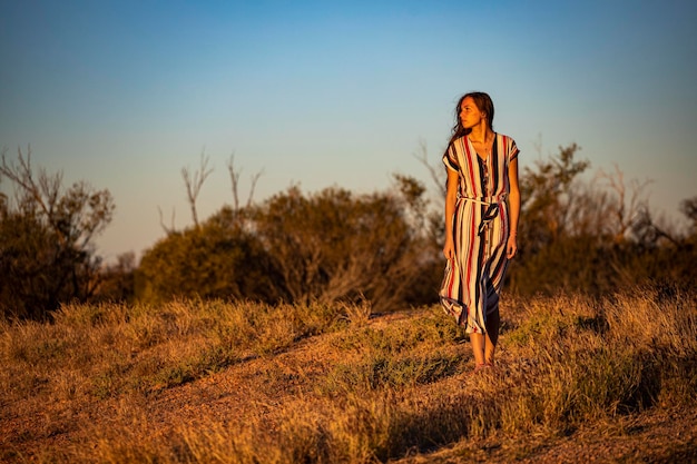 une belle fille aux cheveux longs vêtue d'une robe longue marche le long d'une route dans le désert de l'ouest de l'australie