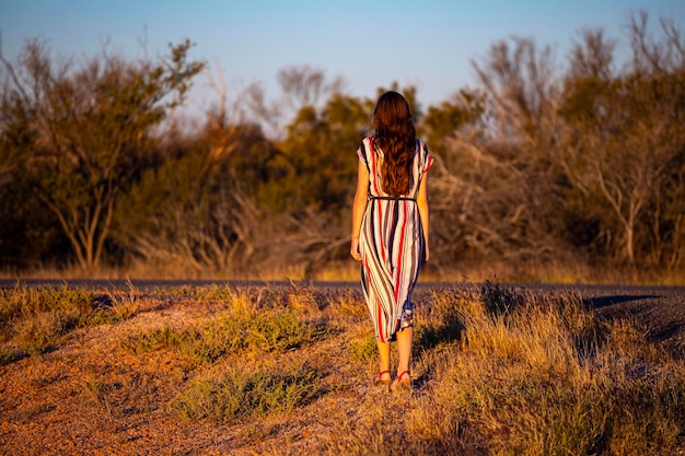 une belle fille aux cheveux longs vêtue d'une robe longue marche le long d'une route dans le désert de l'ouest de l'australie