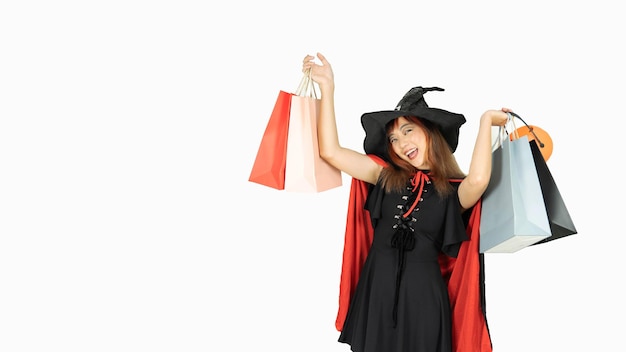 Belle fille aux cheveux noirs et orange en robe noire et chapeau de sorcière tient des sacs à provisions et souriant sur fond blanc pour le jour d'Halloween