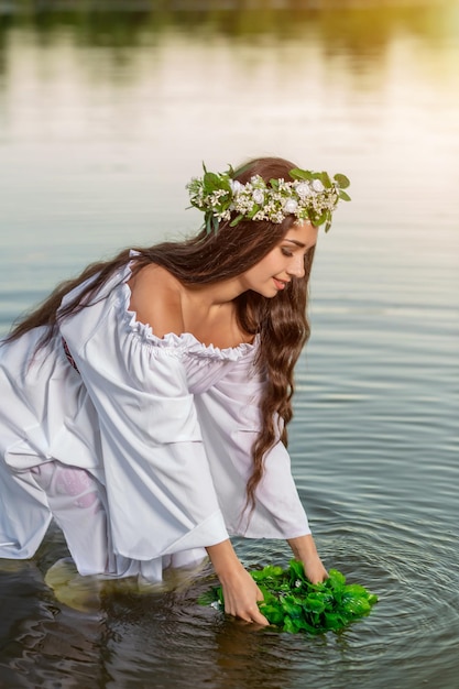 Belle fille aux cheveux noirs en robe vintage blanche et couronne de fleurs debout dans l'eau du lac. Éclat de soleil.