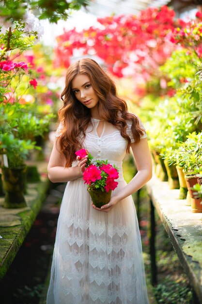 Belle fille aux cheveux rouges dans une robe en dentelle blanche debout dans un jardin avec des fleurs colorées.