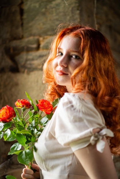 Belle fille aux cheveux roux près de la vieille fenêtre dans une robe de nuit blanche dans une maison traditionnelle française sur le thème de la romance