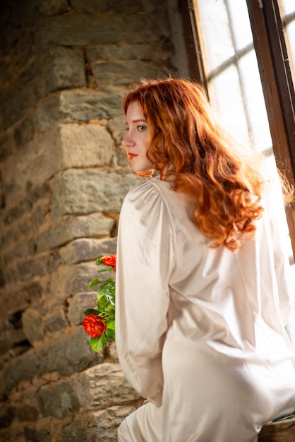 Belle fille aux cheveux roux près de la vieille fenêtre dans une robe de nuit blanche dans une maison traditionnelle française sur le thème de la romance