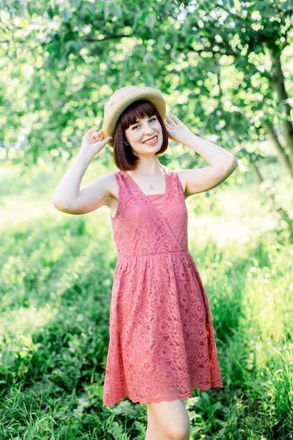 Belle fille aux cheveux roux se dresse sur le fond du jardin de pommes Elle tient son chapeau contre son chapeau et sourit d'un doux sourire. la jeune fille est vêtue d'une robe bleue à pois blancs et d'un chapeau de paille-panama.