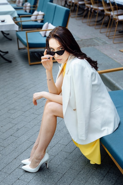 Belle fille aux longues jambes assise au café regarde la caméra sur des lunettes de soleil jeune femme séduisante en jaune