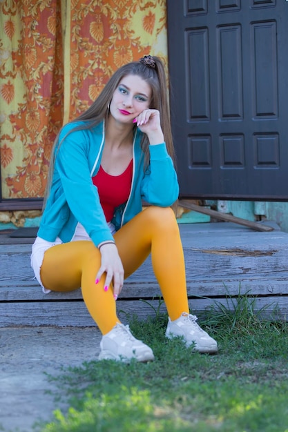 Une belle fille aux vêtements clairs est assise sur le porche d'une maison de village. Femme dans le style des années 90.