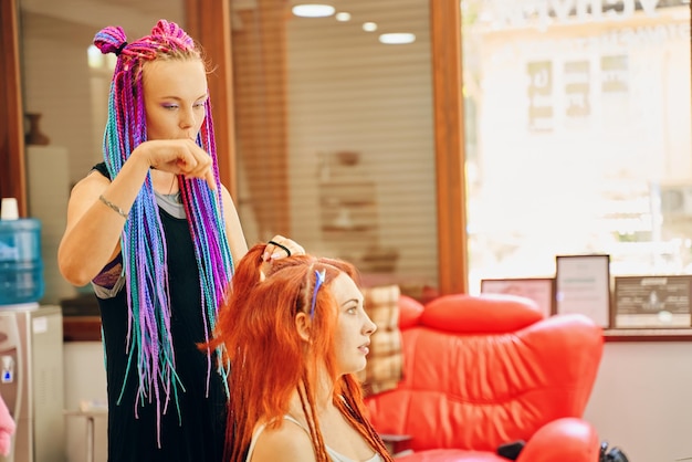 Photo belle fille barbier avec des tresses colorées qui tresse des dreadlocks au gingembre à une femme créative et fashi...