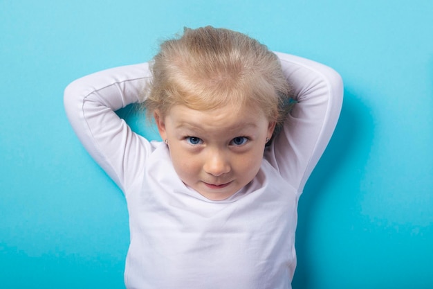 Belle fille blonde allongée tient ses mains derrière sa tête sur un fond bleu. Vue de dessus, mise à plat.