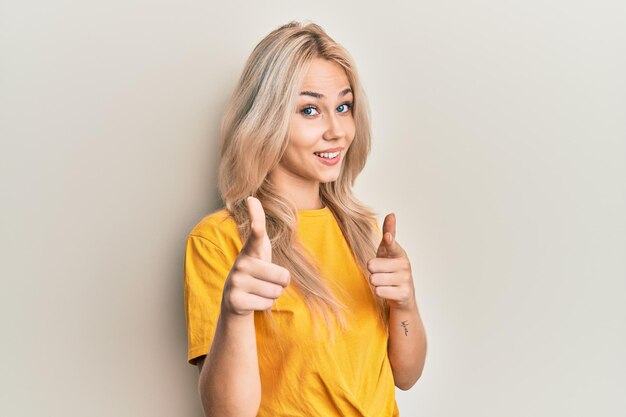 Belle fille blonde caucasienne portant un t-shirt décontracté pointant du doigt l'appareil photo avec un visage heureux et drôle, une bonne énergie et des vibrations