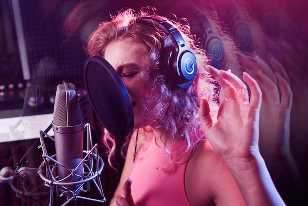 Belle fille blonde chantant émotionnellement une chanson dans un studio d'enregistrement avec un microphone et un casque professionnels crée un nouvel album de chansons artiste vocal en néon bleu rose face agrandi