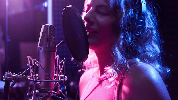 Belle fille blonde chantant émotionnellement une chanson en studio d'enregistrement avec microphone professionnel et écouteurs, crée un nouvel album de piste, artiste vocal en néon bleu rose, visage en gros plan