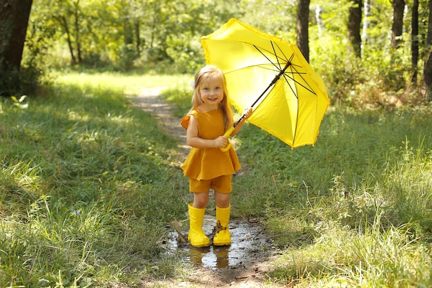 une belle fille blonde dans un costume de lin en bottes jaunes avec un parapluie jaune se dresse dans le parc
