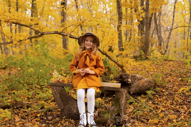 une belle fille blonde dans un manteau beige et un chapeau est assise sur un banc dans le parc de la ville en automne