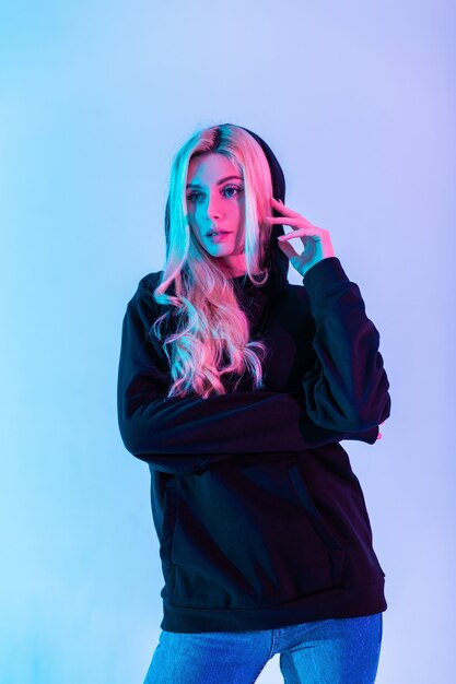 Belle fille blonde glamour dans un sweat à capuche noir à la mode avec un jean posant en studio sur un fond rose néon coloré