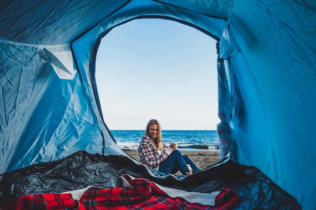 Belle fille blonde joyeuse souriante vue de l'intérieur d'une tente campée sur la plage directement sur le sable