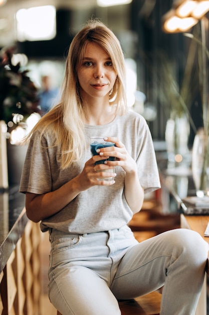 Une belle fille blonde mince, vêtue d'un style décontracté, est assise sur la chaise et tient une tasse de café dans un café confortable. .