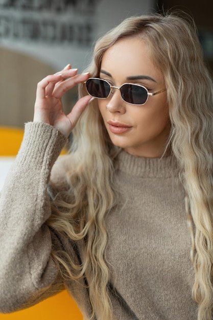 Belle fille blonde à la mode dans des vêtements à la mode avec un pull beige met des lunettes de soleil vintage et s'assied dans un café près de la fenêtre